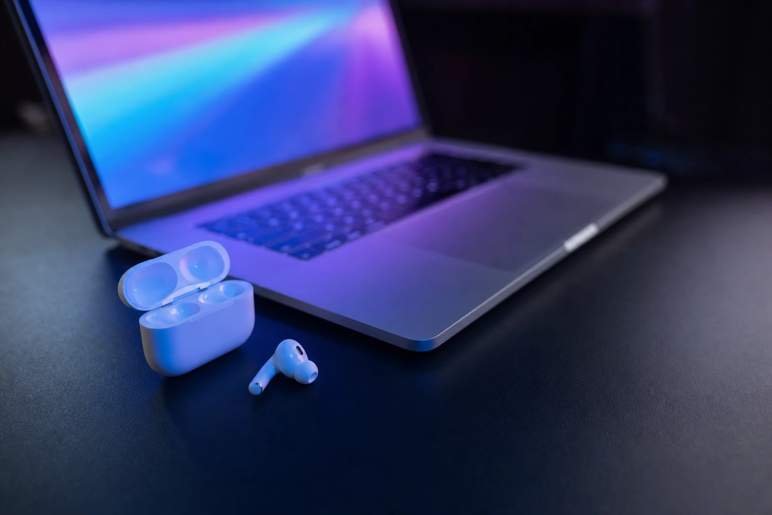 White earbud and open case on dark slate, lit by blue and violet lights. A blurred, glowing laptop is in the background.