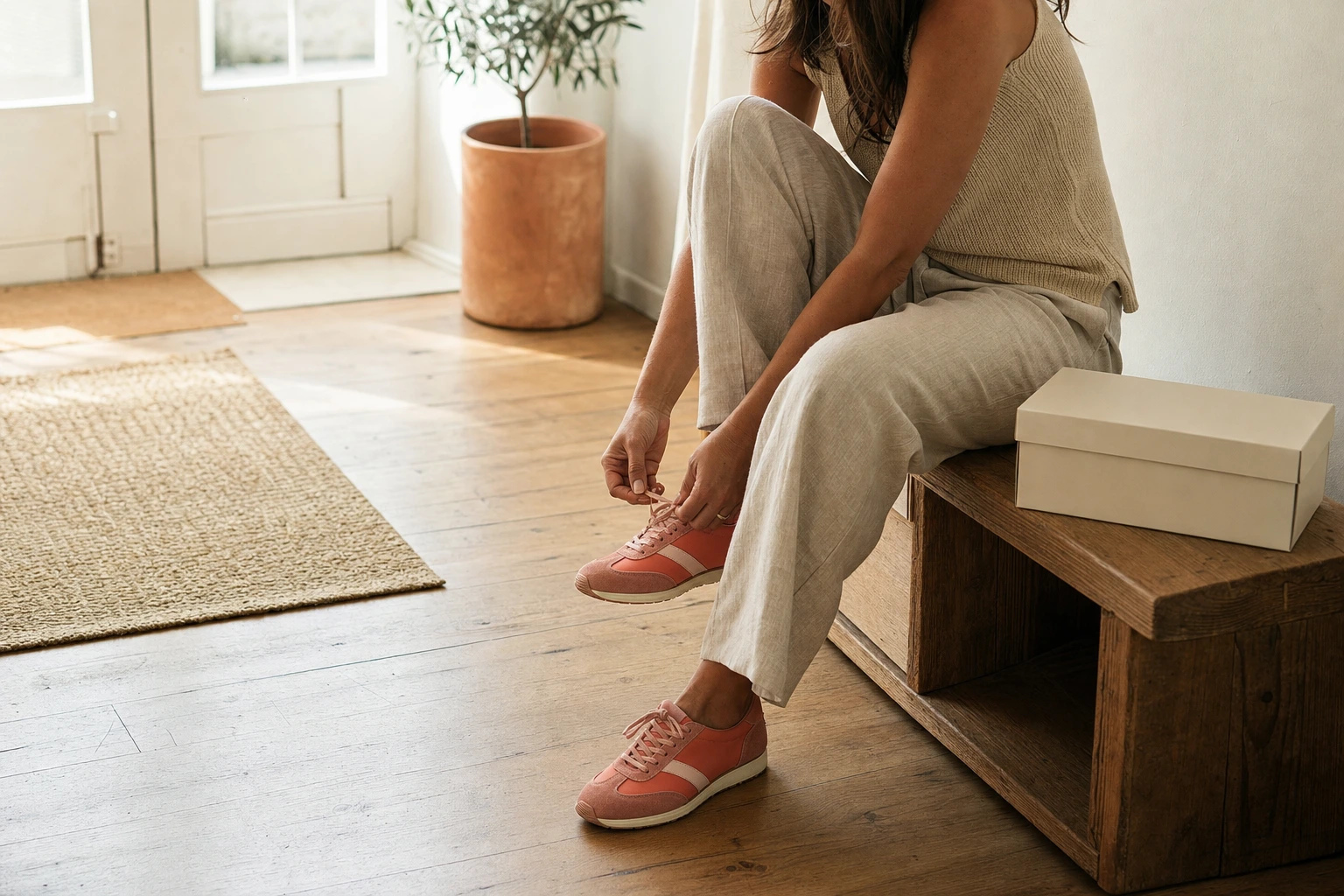 A woman on a wooden bench in a sunlit entryway laces pink sneakers, wearing linen trousers. A plain shoebox sits on the floor.